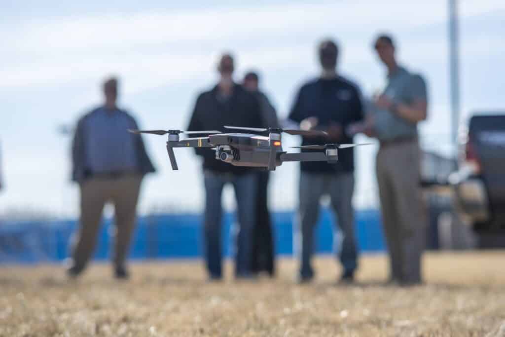 A drone hovers close to the ground during an outdoor demonstration, with a group of people standing in the blurred background watching the flight.