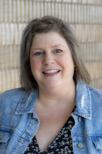 Professional headshot of a woman with shoulder-length brown hair wearing a denim jacket over a patterned blouse, smiling in front of a light brick wall background.