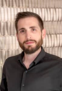 Professional headshot of a man with short brown hair and a trimmed beard wearing a black button-up shirt, in front of a softly blurred neutral background.