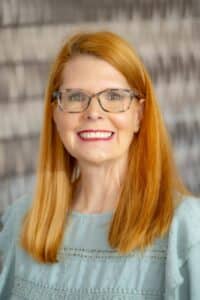 Professional headshot of a smiling woman with straight red hair and glasses, wearing a light blue blouse with lace details, in front of a softly blurred neutral background.