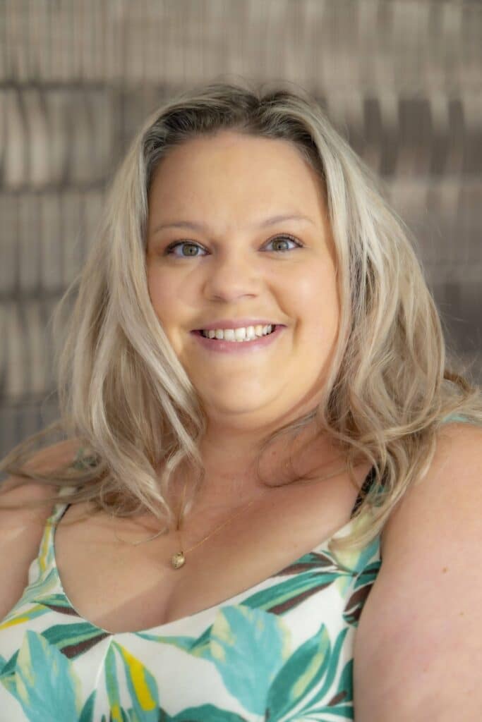 Professional headshot of a smiling woman with blonde hair, wearing a white and green patterned top, in front of a softly blurred neutral background.