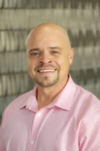 Professional headshot of a smiling man with a shaved head and goatee, wearing a light pink collared shirt, in front of a softly blurred neutral background.