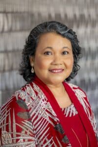 Professional headshot of a smiling woman with short curly salt-and-pepper hair, wearing a red patterned blouse and matching shawl, in front of a softly blurred neutral background.