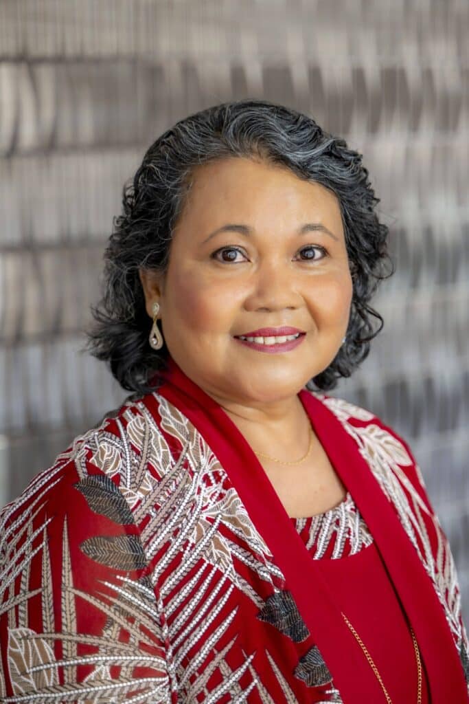 Professional headshot of a smiling woman with short curly salt-and-pepper hair, wearing a red patterned blouse and matching shawl, in front of a softly blurred neutral background.