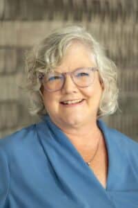 Professional headshot of a smiling woman with short curly gray hair and glasses, wearing a light blue blouse, in front of a softly blurred neutral background.