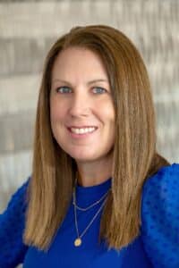 Professional headshot of a smiling woman with straight brown hair wearing a royal blue blouse and gold layered necklaces, in front of a softly blurred neutral background.