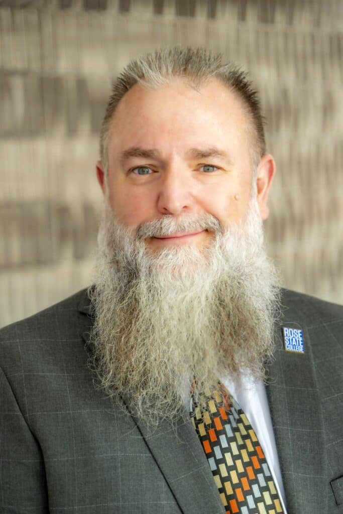 Professional headshot of a smiling man with a long gray beard and short hair, wearing a gray suit jacket, patterned tie, and a Rose State College lapel pin, in front of a softly blurred neutral background.
