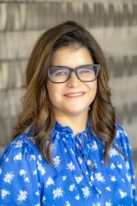 Professional headshot of a smiling woman with long brown hair and glasses wearing a royal blue floral blouse, in front of a softly blurred neutral background.