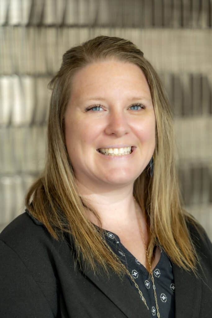 Professional headshot of a smiling woman with straight blonde hair wearing a black blazer and navy patterned blouse, in front of a softly blurred neutral background.