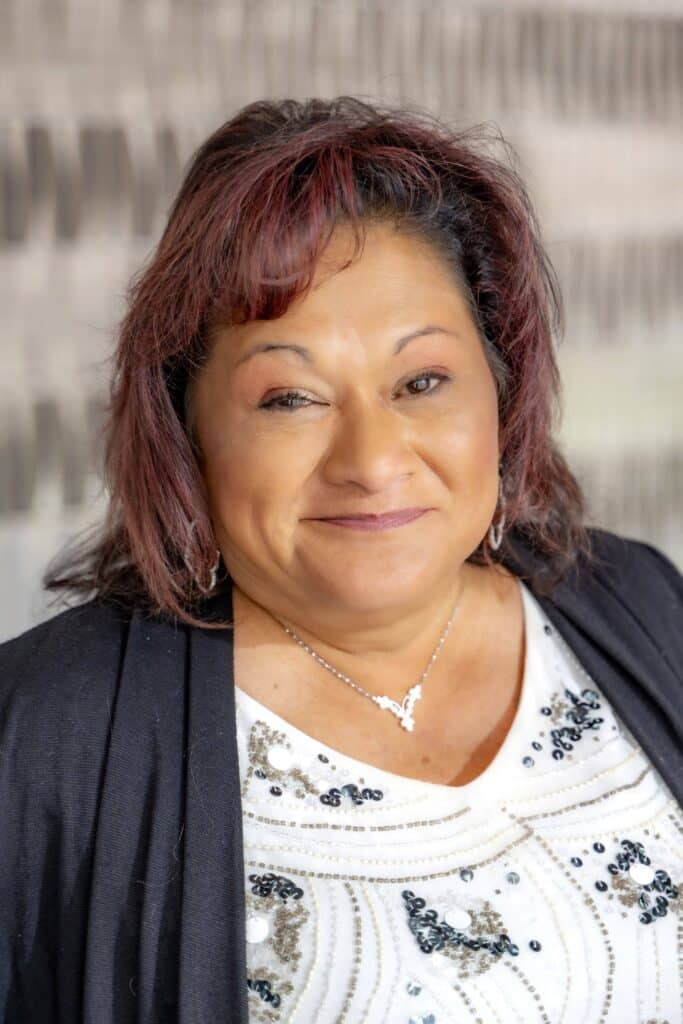 Professional headshot of a smiling woman with shoulder-length dark red hair wearing a white top with black and silver beading and a black cardigan, in front of a softly blurred neutral background.