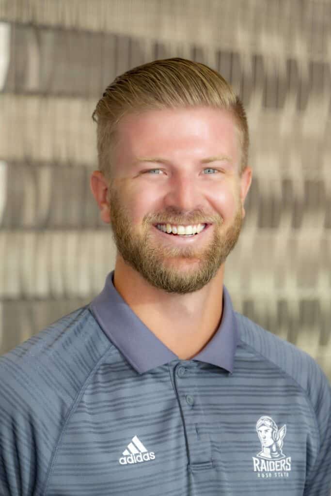 Professional headshot of a smiling man with short light brown hair and beard wearing a gray Adidas Rose State Raiders polo shirt, in front of a softly blurred neutral background.
