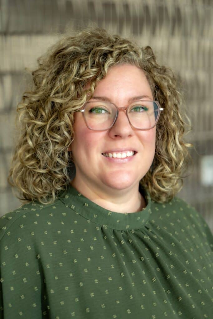 Professional headshot of a smiling woman with curly blonde hair and glasses wearing a green blouse with a subtle dotted pattern, in front of a softly blurred neutral background.