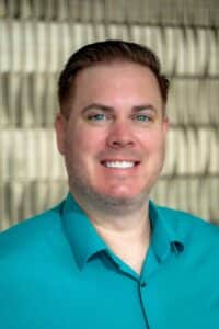 Professional headshot of a smiling man wearing a teal button-up shirt, in front of a softly blurred neutral background.
