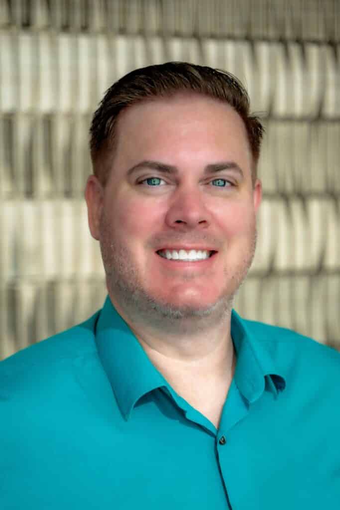 Professional headshot of a smiling man wearing a teal button-up shirt, in front of a softly blurred neutral background.