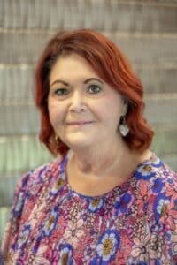 Professional headshot of a woman with short red hair wearing a colorful floral blouse and heart-shaped earrings, in front of a softly blurred neutral background.