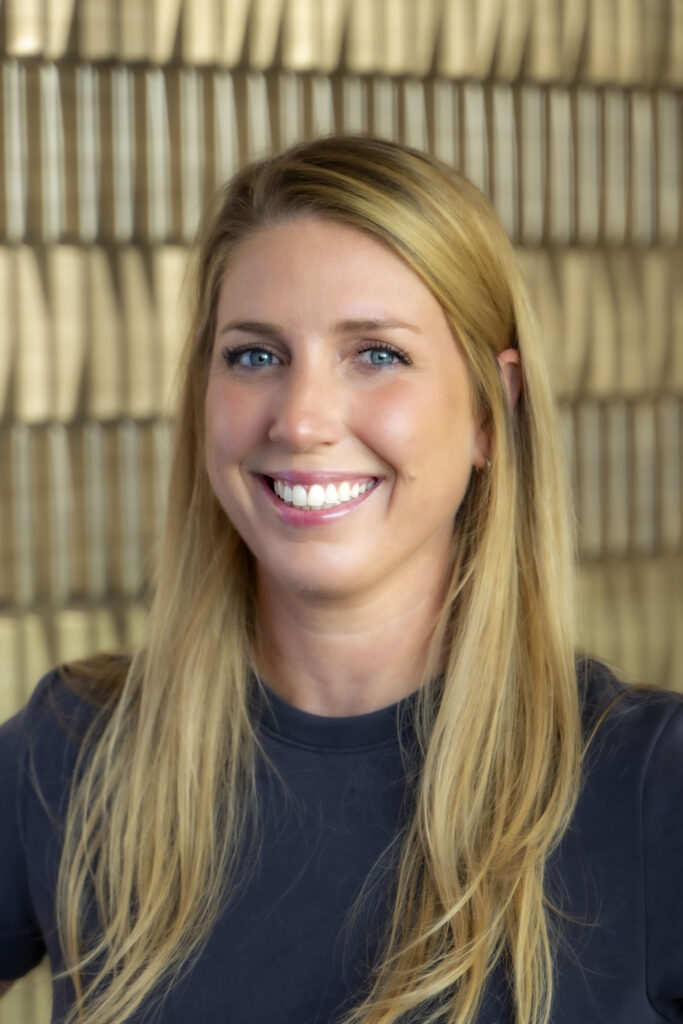 Smiling woman with long blonde hair wearing a dark top against a softly blurred neutral background.