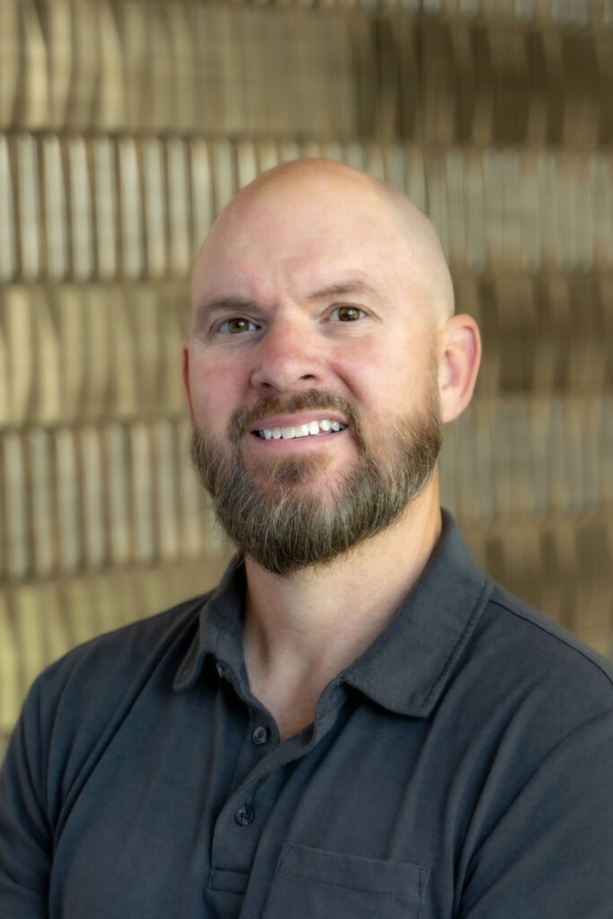 Smiling bald man with a full beard wearing a dark gray collared shirt.