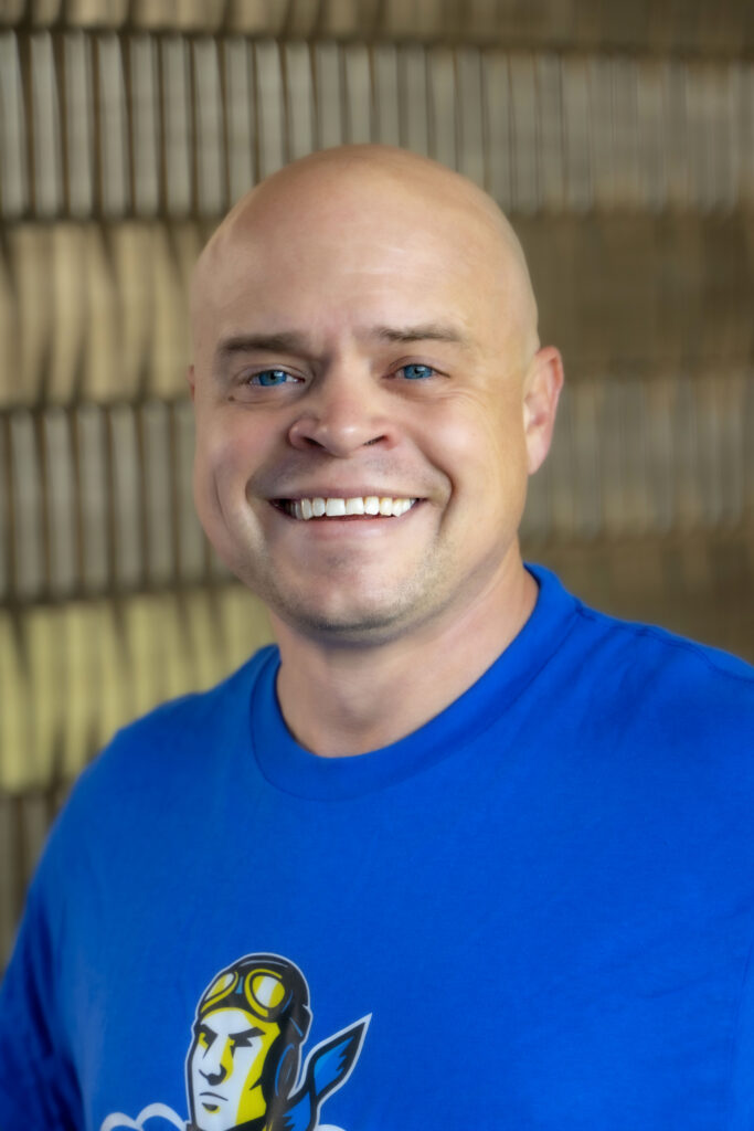 Professional headshot of a smiling person with shoulder-length dark hair wearing a black top against a softly blurred neutral background.