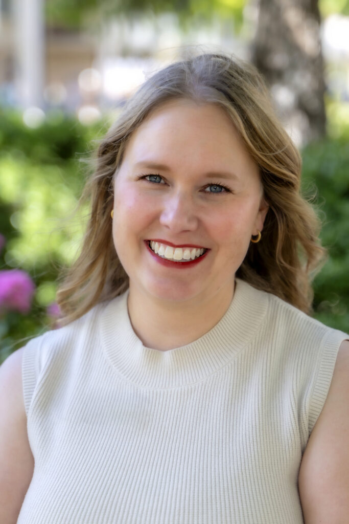 Professional headshot of a smiling woman with shoulder-length light brown hair wearing a sleeveless cream knit top, photographed outdoors with trees and greenery softly blurred in the background.