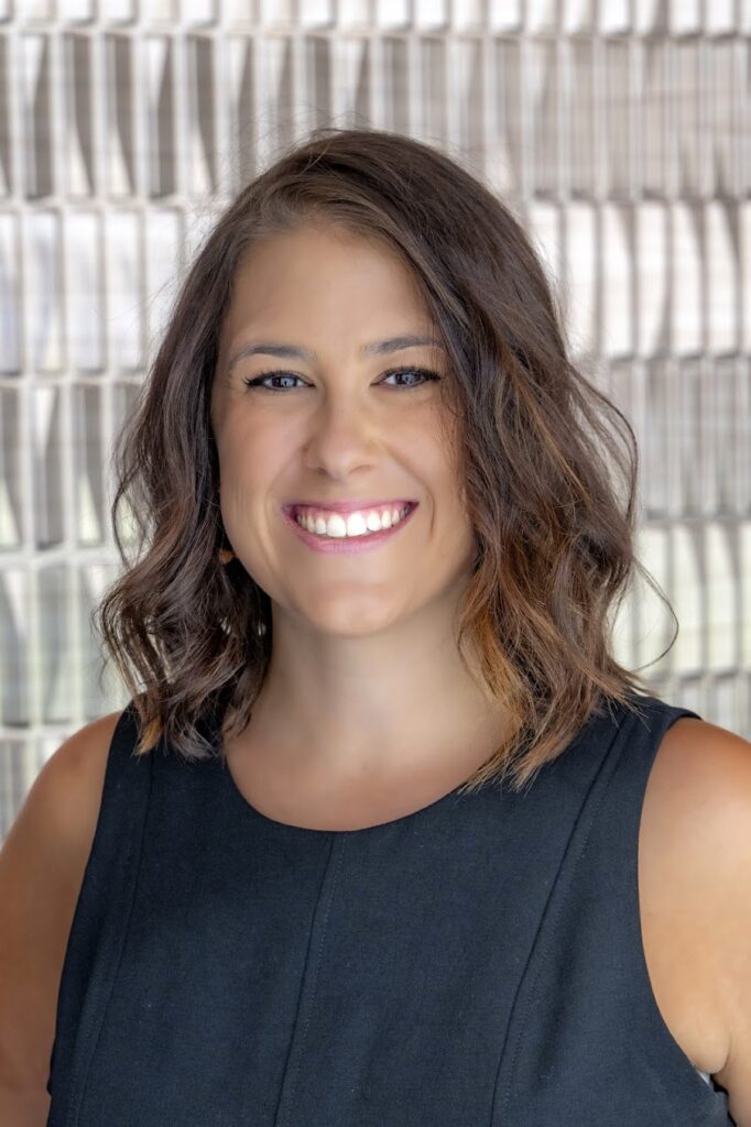 Portrait of a smiling woman with shoulder-length wavy brown hair, wearing a sleeveless black top, standing in front of a softly patterned background.