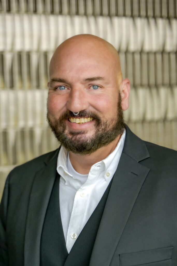 Portrait of a smiling man with a beard wearing a dark suit jacket and white shirt, standing in front of a softly blurred indoor background.