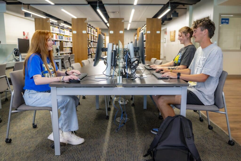 three students sit typing on computers at a table in the library.