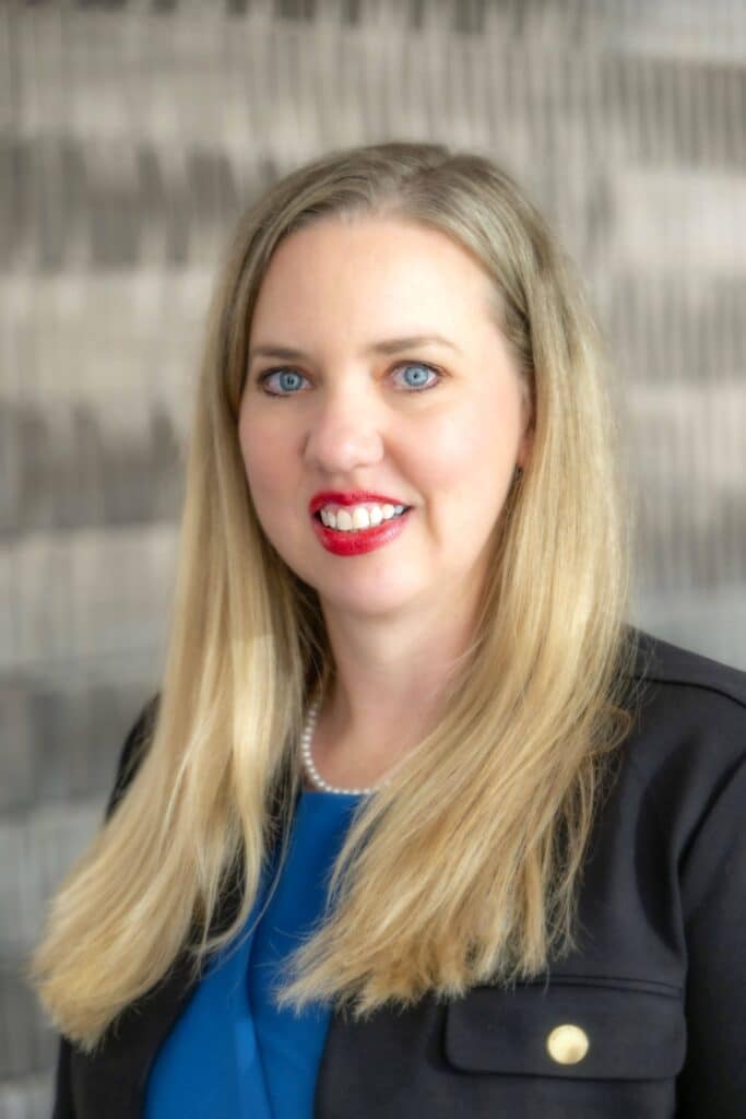 Professional headshot of a woman with long blonde hair wearing a blue blouse, black blazer, pearl necklace, and red lipstick, in front of a softly blurred neutral background.