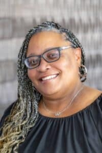 Professional headshot of a smiling woman with braided hair wearing glasses, hoop earrings, and a black blouse, in front of a softly blurred neutral background.