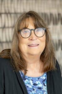 Professional headshot of a woman with shoulder-length brown hair wearing glasses, a black cardigan, and a blue floral blouse, in front of a softly blurred neutral background.