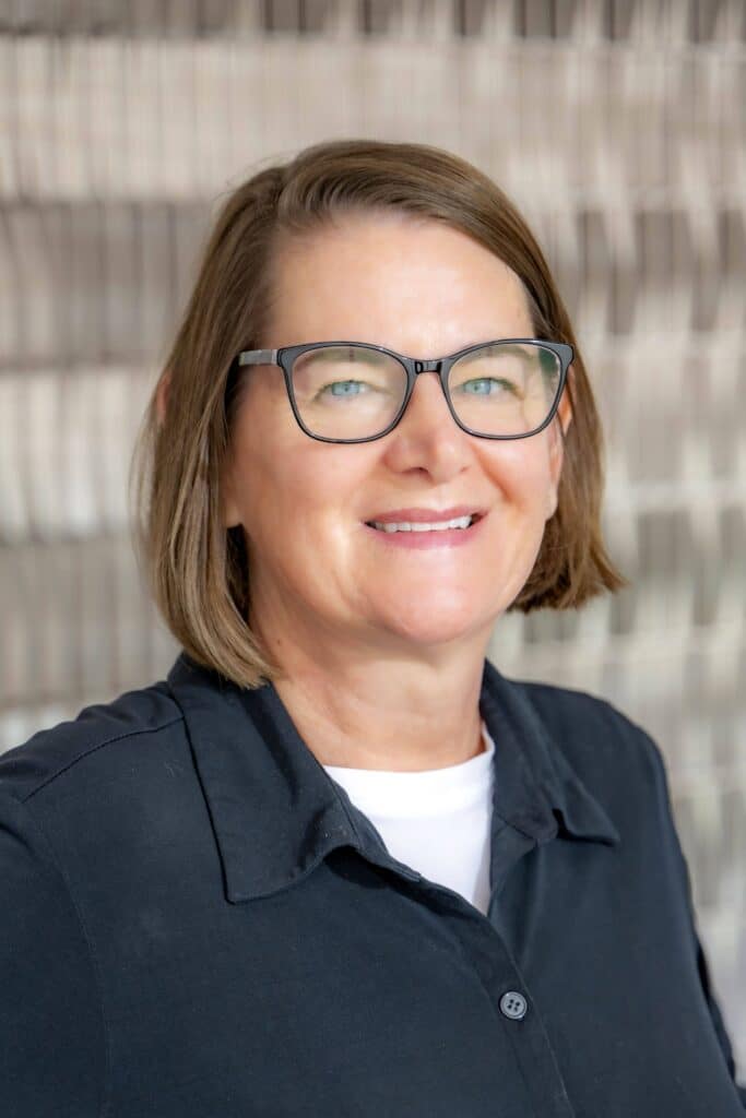 Professional headshot of a smiling woman with short brown hair wearing glasses, a black collared shirt, and a white undershirt, in front of a softly blurred neutral background.