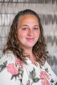 Professional headshot of a smiling woman with wavy brown hair wearing a white blouse with a pink floral pattern, posed in front of a softly blurred neutral background.