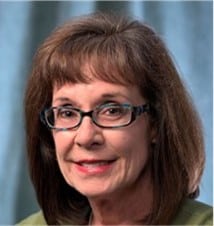 Professional headshot of a smiling woman with shoulder-length brown hair and glasses, wearing a green top, in front of a light blue background.