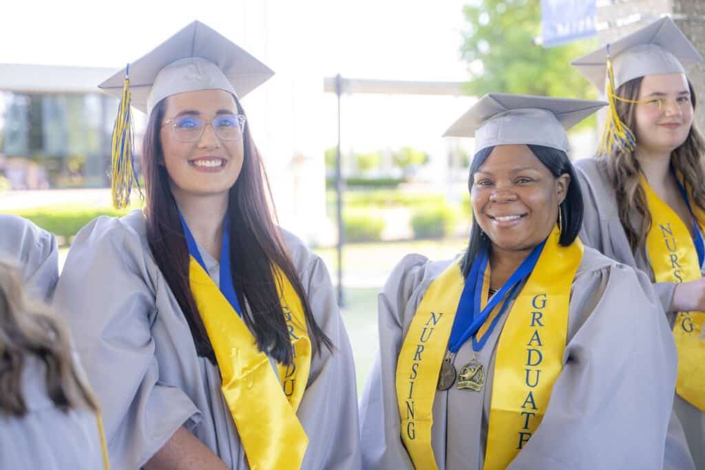Three nursing graduates wearing gray caps and gowns with yellow graduation stoles smile while standing together outside before the ceremony.