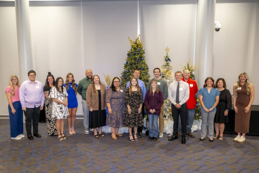 A large group of people standing in a row and smiling in front of decorated Christmas trees at an indoor Rose State College event.