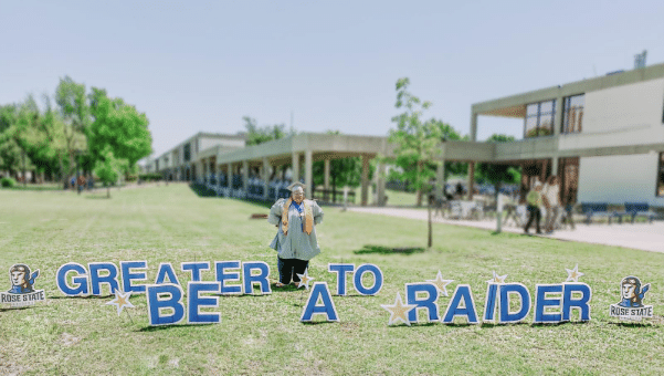 A graduate in cap and gown stands on a grassy lawn at Rose State College behind large blue yard letters that read 'BE A GREATER RAIDER,' with campus buildings visible in the background.