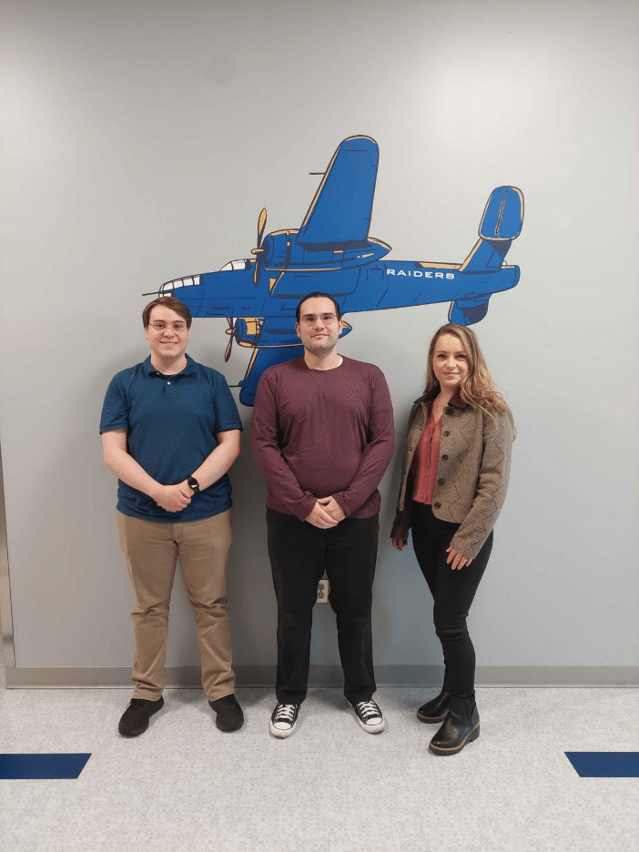 Three people standing and smiling in front of a wall mural of a blue airplane with the word 'Raiders' on it inside a Rose State College building.