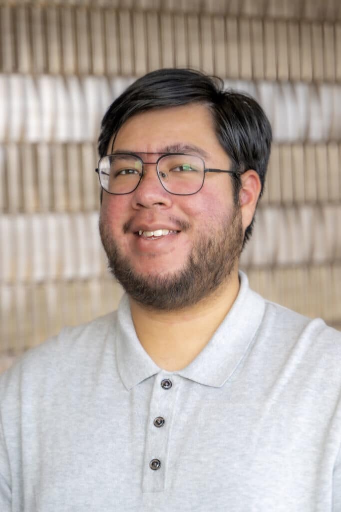 Portrait of a smiling man wearing glasses and a light gray collared shirt, standing in front of a softly blurred patterned background.