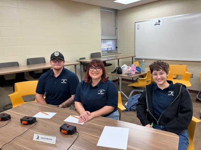 Three students wearing matching navy shirts sit at a table with quiz buzzers and paper in a classroom.