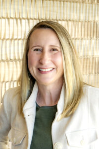 Portrait of a smiling woman with blonde hair wearing a cream jacket over a green top, photographed against a light textured background.