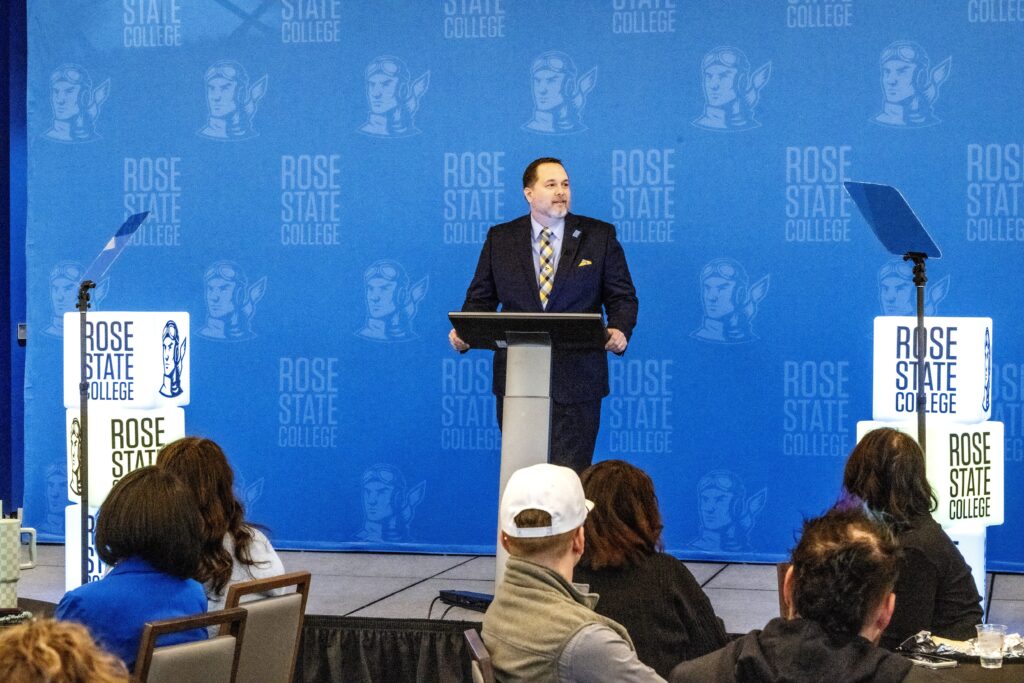 Speaker at a podium addresses an audience in front of a blue Rose State College backdrop, with teleprompters on both sides of the stage.