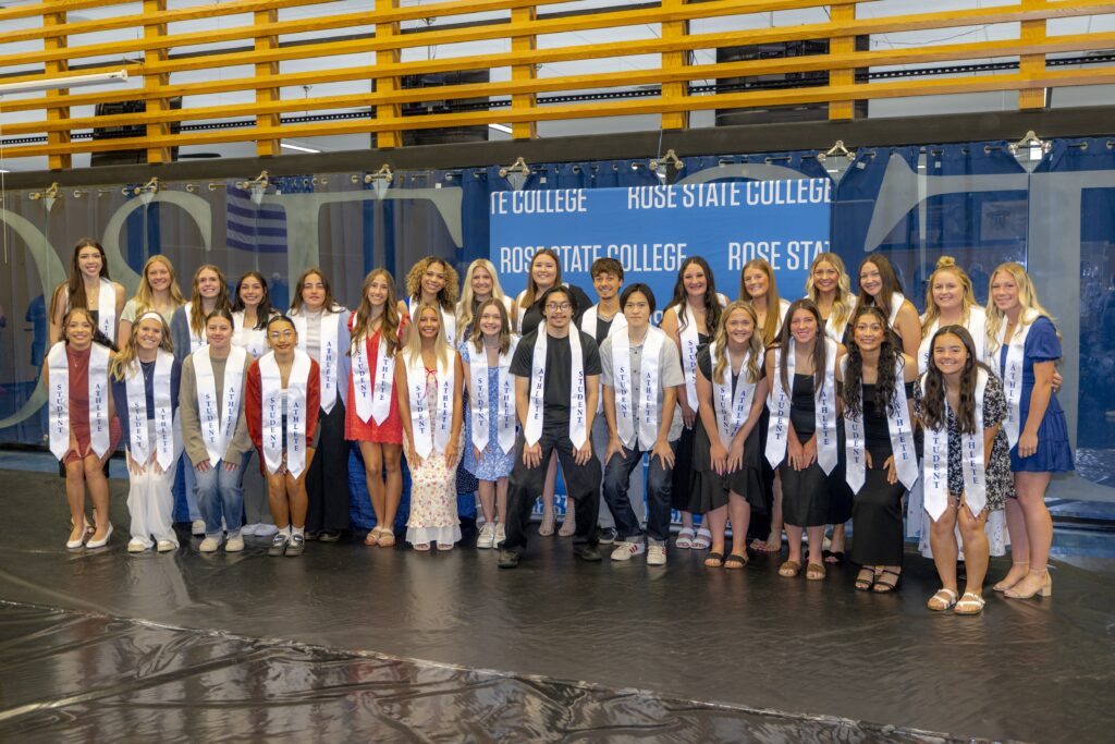 Rose State College student-athletes pose for a group photo in front of a Rose State College backdrop, wearing white stoles at an athletics graduation ceremony.