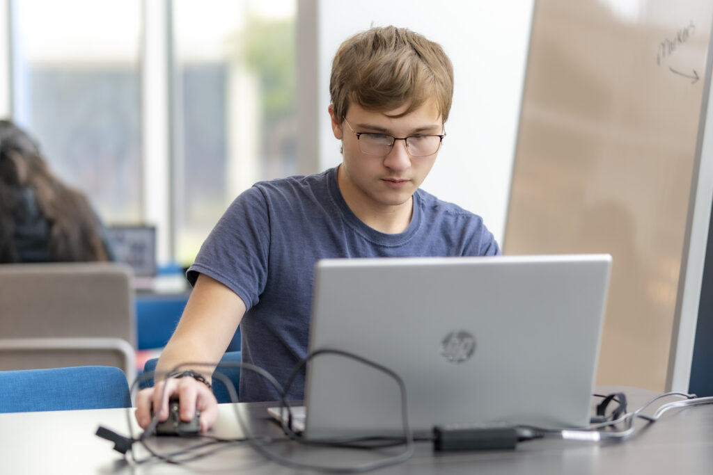 Student works on a laptop at a table in a campus study area, using a computer mouse and focusing on the screen.