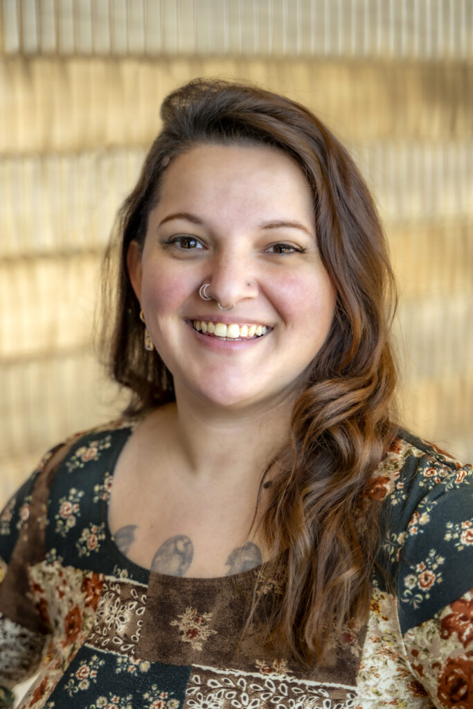 Portrait of a smiling woman with long wavy hair, wearing a patterned top, photographed indoors against a softly blurred background.