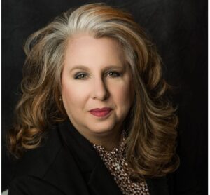 Professional headshot of a woman with shoulder-length styled hair wearing a black blazer and patterned blouse, posed against a dark background.