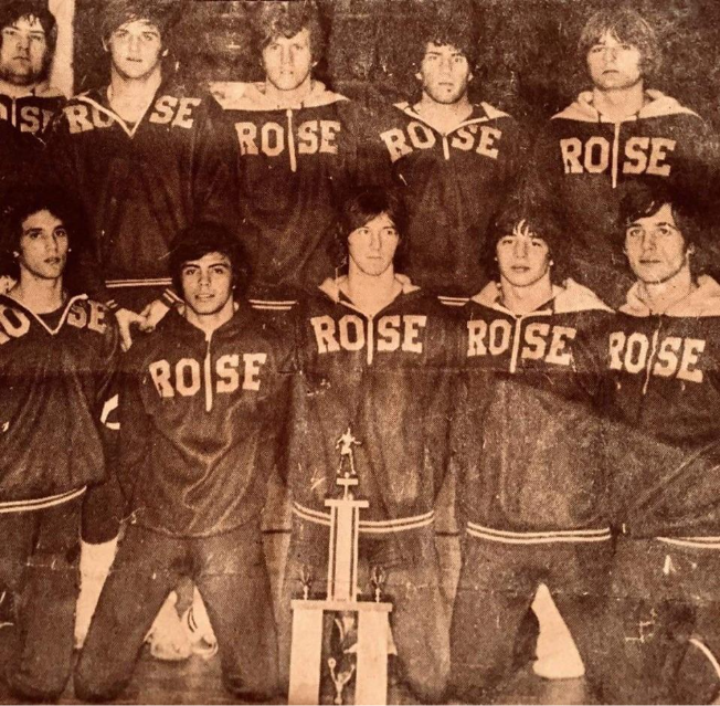 Vintage black-and-white team photo of Rose basketball players wearing “ROSE” warm-up jackets, posed in two rows with a trophy displayed in front.