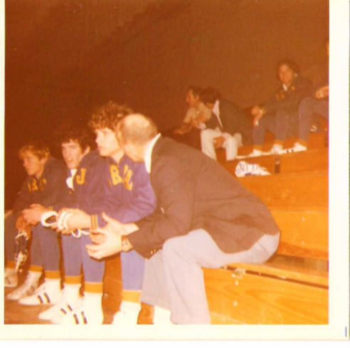 Vintage color photo of a wrestling coach speaking with players seated on wooden gym bleachers during a game, with other team members visible in the background.