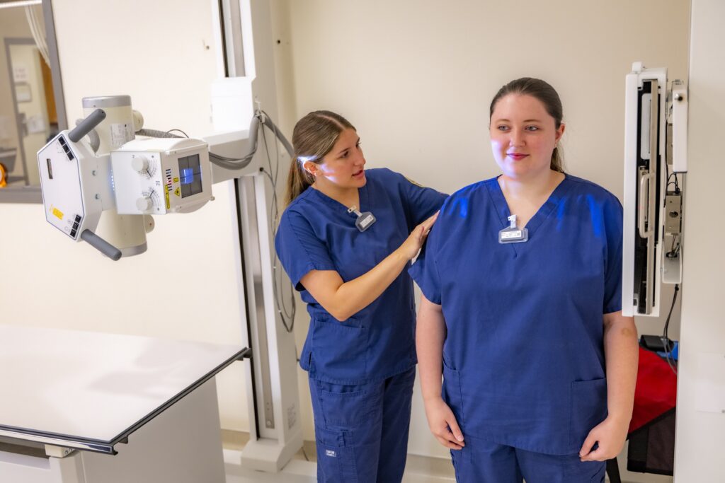 Two students in blue medical scrubs practice patient positioning with X-ray equipment in a radiologic technology lab.