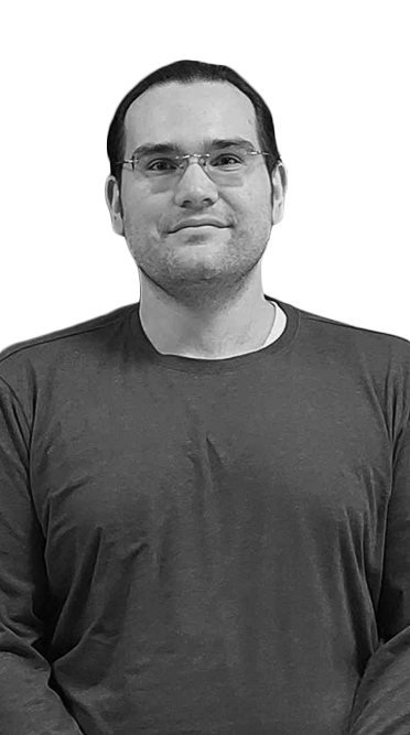 Black-and-white portrait of a man with glasses and short dark hair wearing a long-sleeve shirt, standing and looking at the camera.