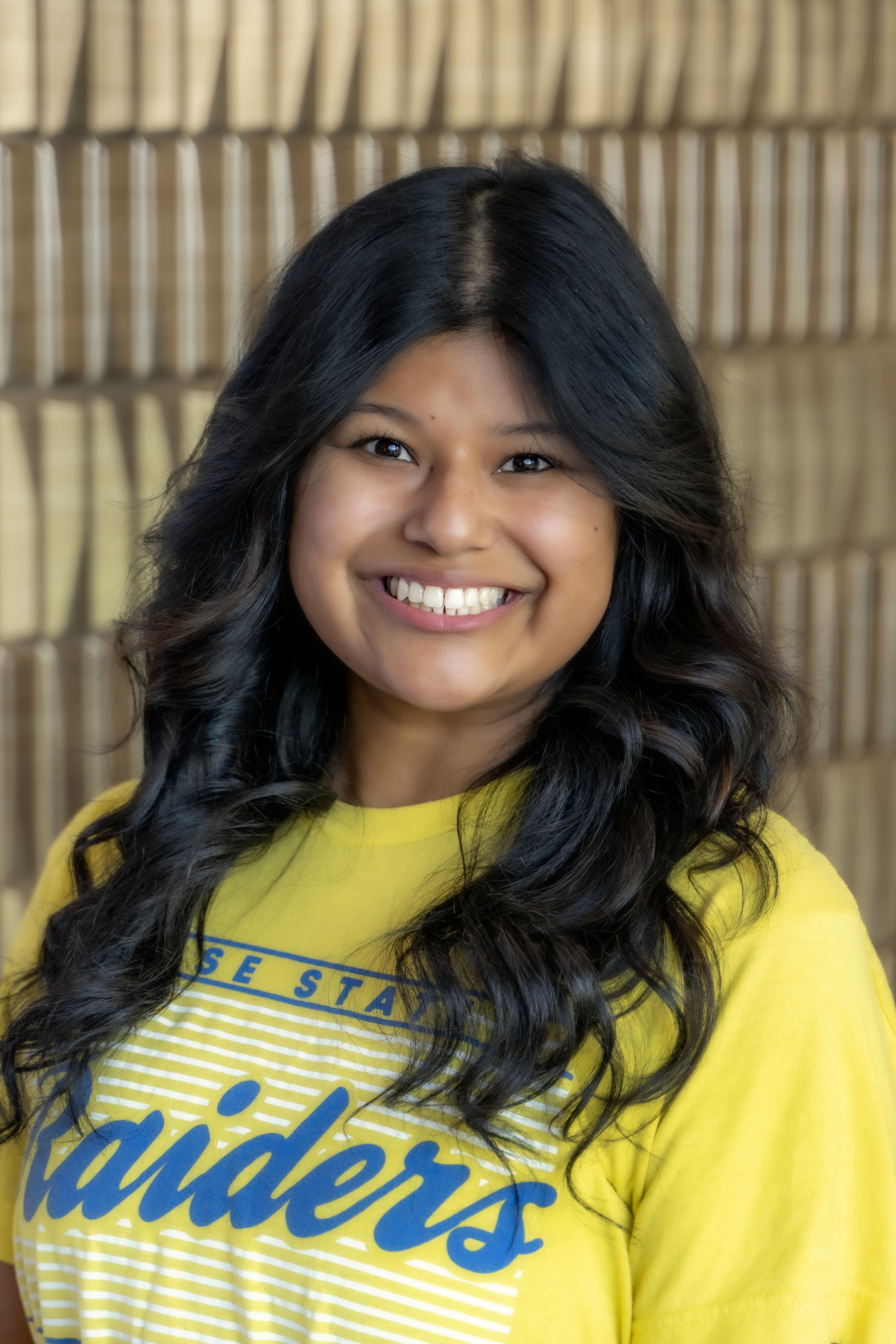 Smiling woman with long dark hair wearing a yellow Rose State Raiders T-shirt against a softly blurred neutral background.