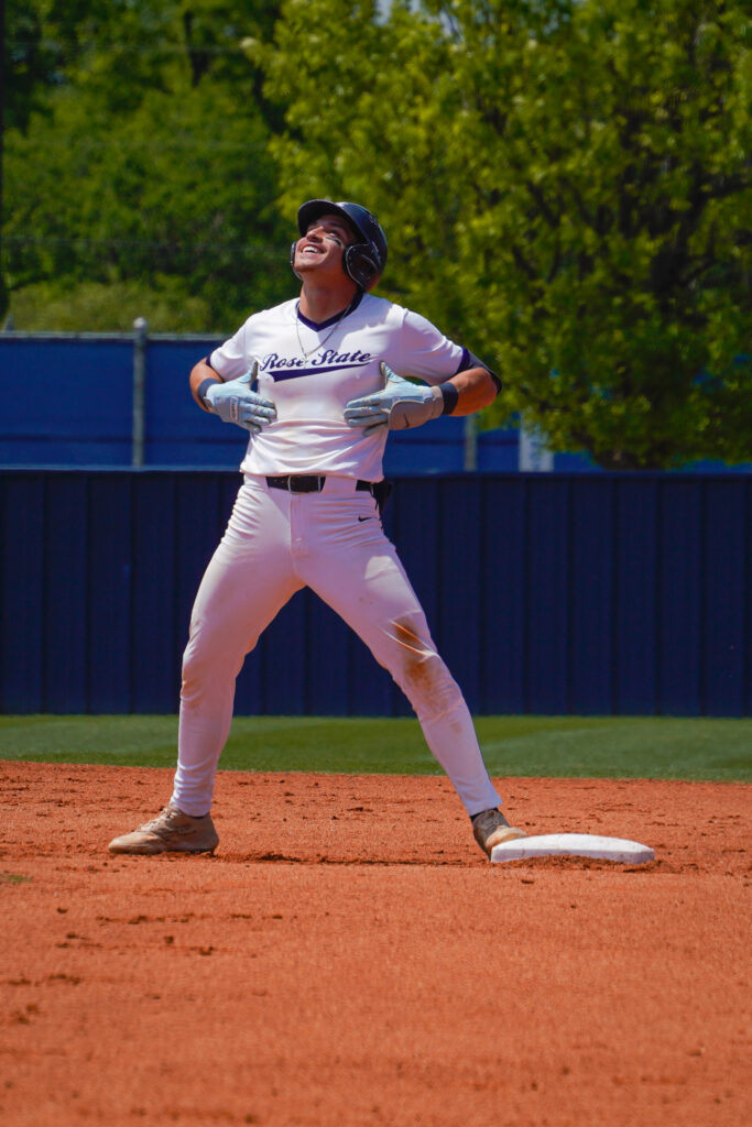 Rose State College baseball player celebrates while standing on base during a game on a sunny day.